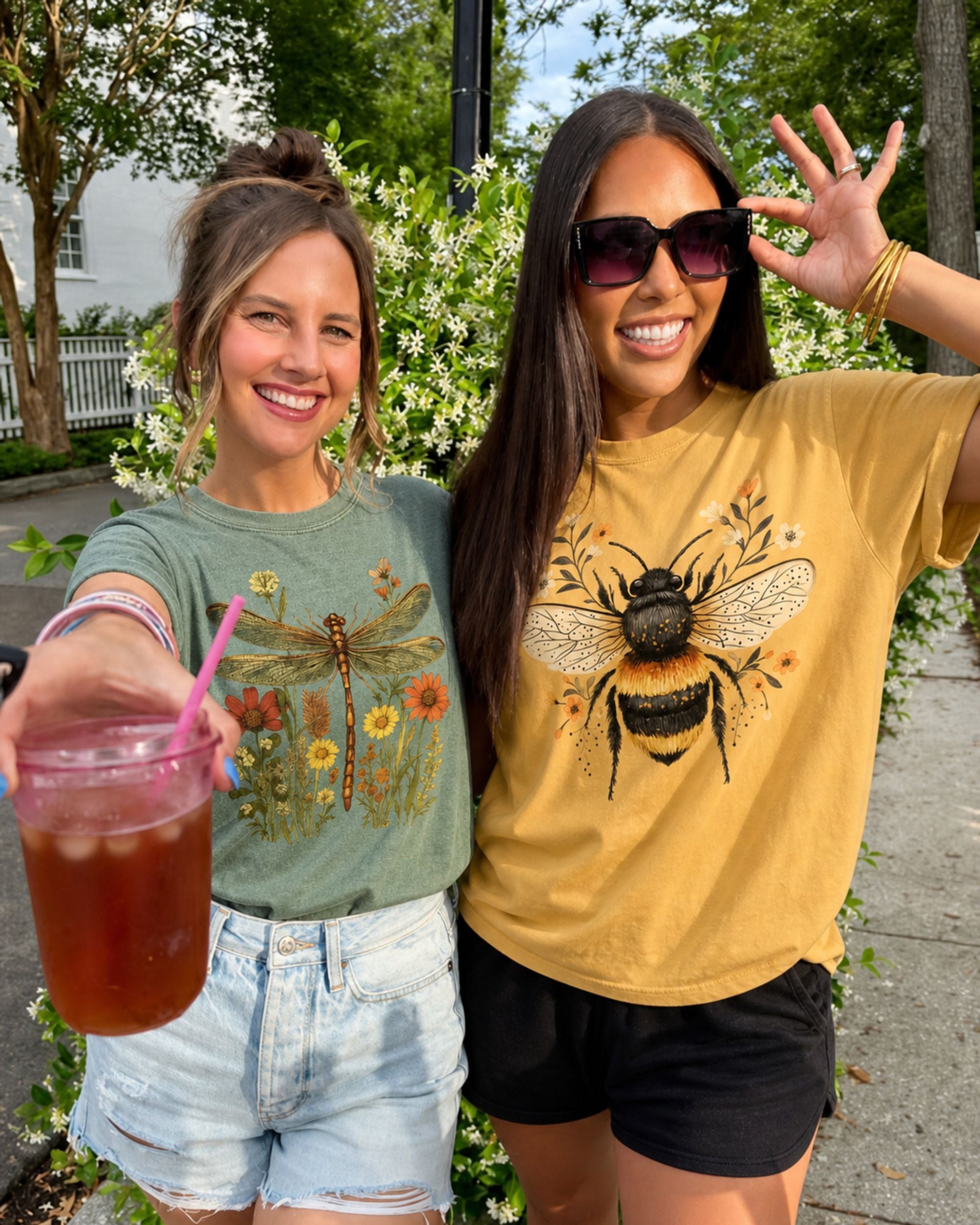 Two women standing outdoors, one holding a drink, both wearing t-shirts with nature-themed designs.