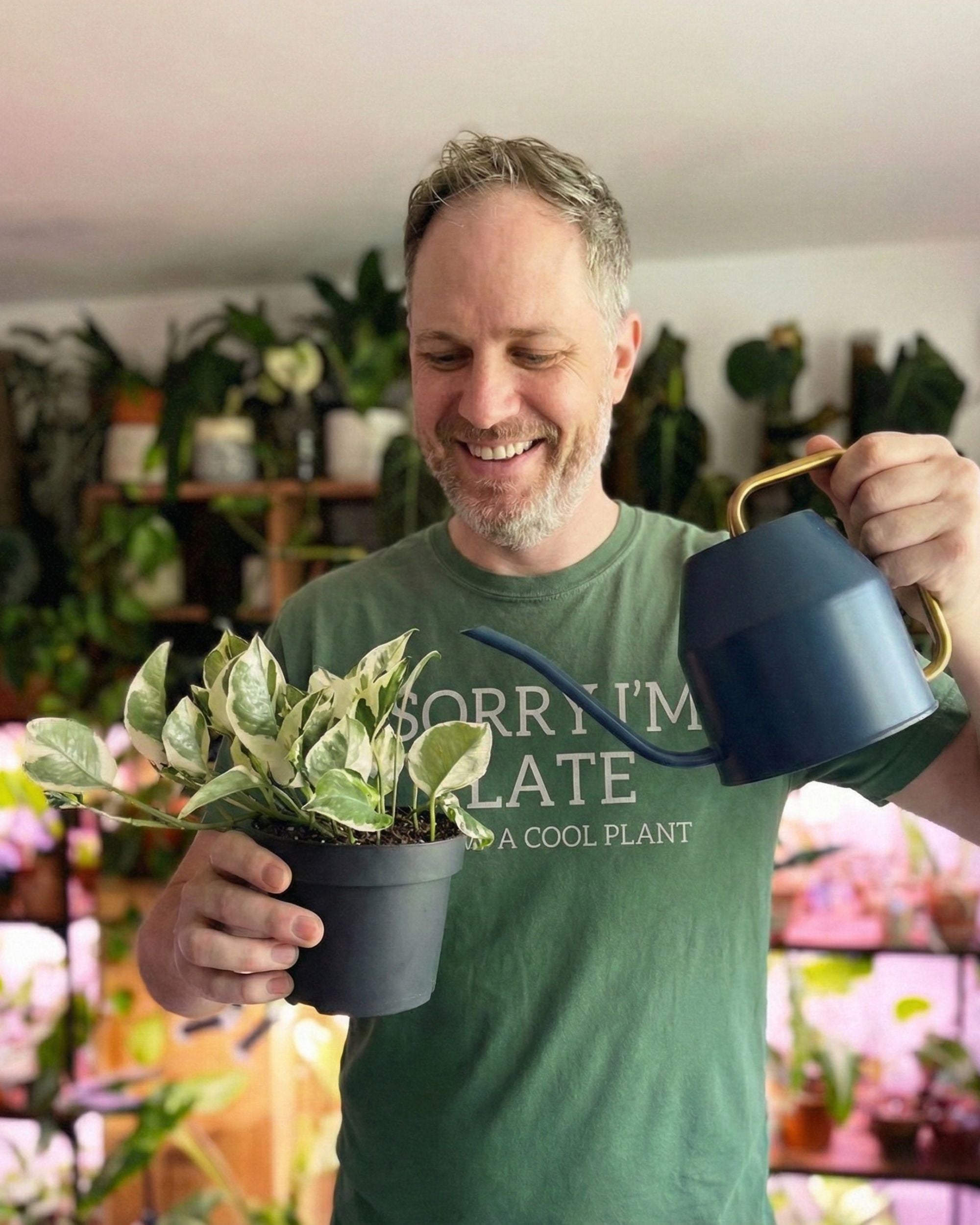 Man holding a potted plant and a watering can in a greenhouse setting.